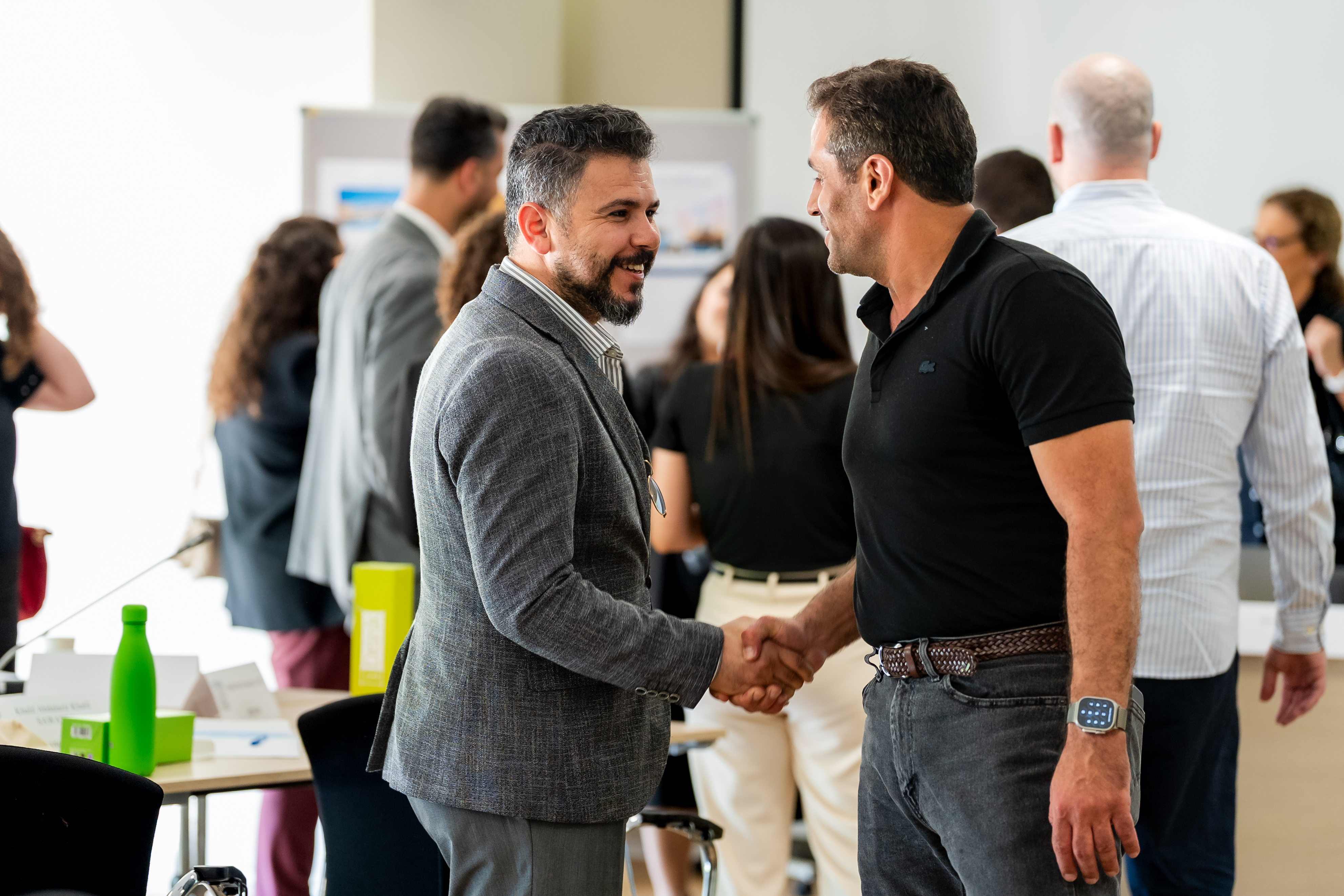 Two programme participants shake hands and smile during a networking moment, with other participants conversing in the background.