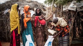 A group of three women, a baby, a toddler and a man gather outside a rustic shelter made of branches and fabric. One woman carries the infant in a bright red cloth sling, while the others wear colorful  yellow and orange headscarves, creating a warm scene of everyday community life.  