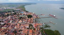 Vista aérea panorâmica de uma cidade portuária cheia de vida, onde os edifícios se encontram com um porto repleto de contentores e longos cais. Os navios de carga repousam na baía tranquila, enquanto mangais exuberantes e zonas húmidas contornam a linha costeira sob um céu enevoado.