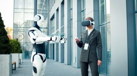 White robot interacts with female worker wearing virtual reality goggles and a grey suit