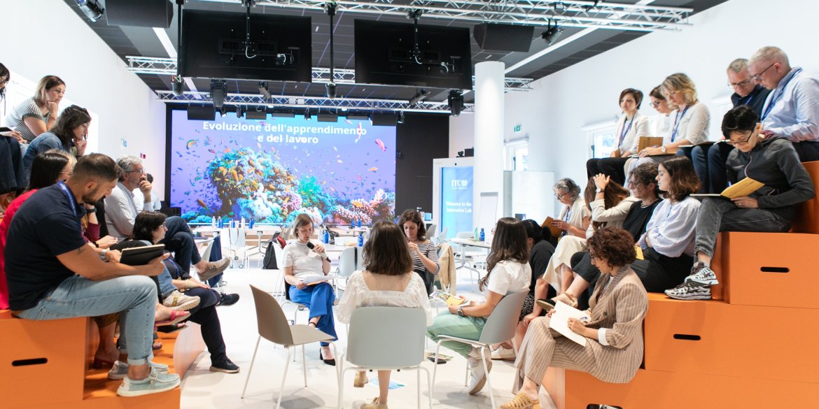 Participants seated in small groups and on tiered seating during an interactive workshop in a modern learning space at the ITCILO campus.