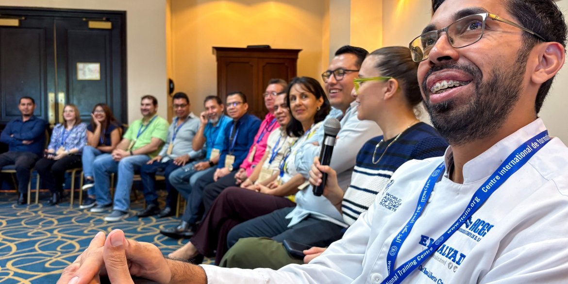 rogramme participants seated in a circle during an interactive group discussion, with one participant speaking into a microphone as others listen and smile.