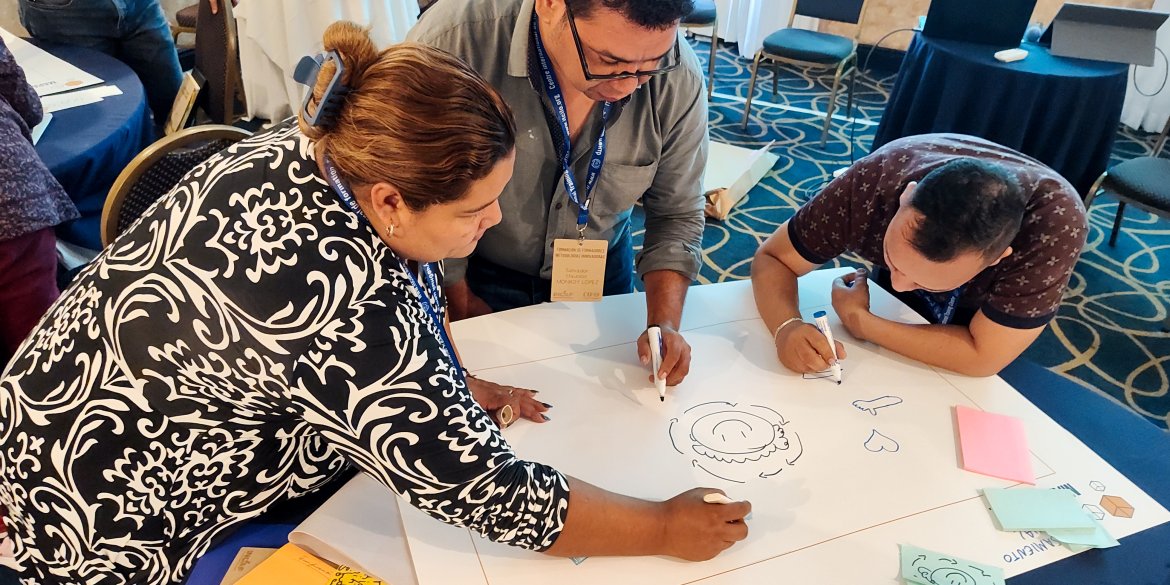 Three participants collaborate around a table, drawing and writing ideas on a large sheet of paper during a hands-on group exercise.