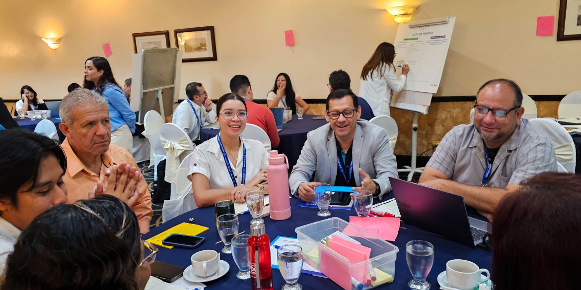 Participants seated around a table engage in a lively group discussion during a workshop, while other groups work on activities in the background.