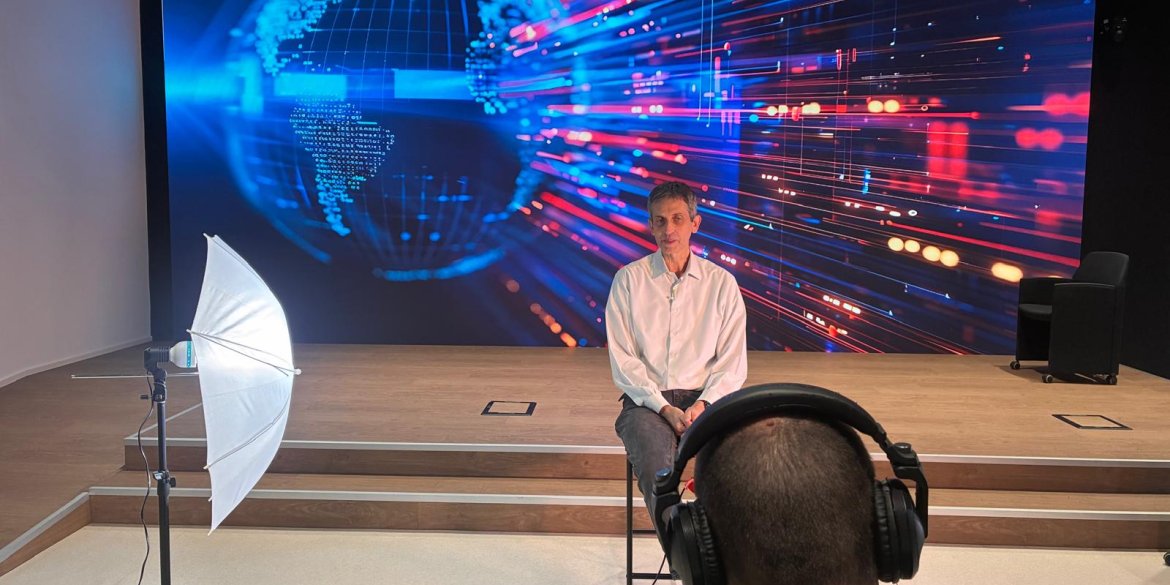 A behind-the-scenes view of a video interview, with a man seated in front of a colourful digital globe backdrop as a crew member records him.