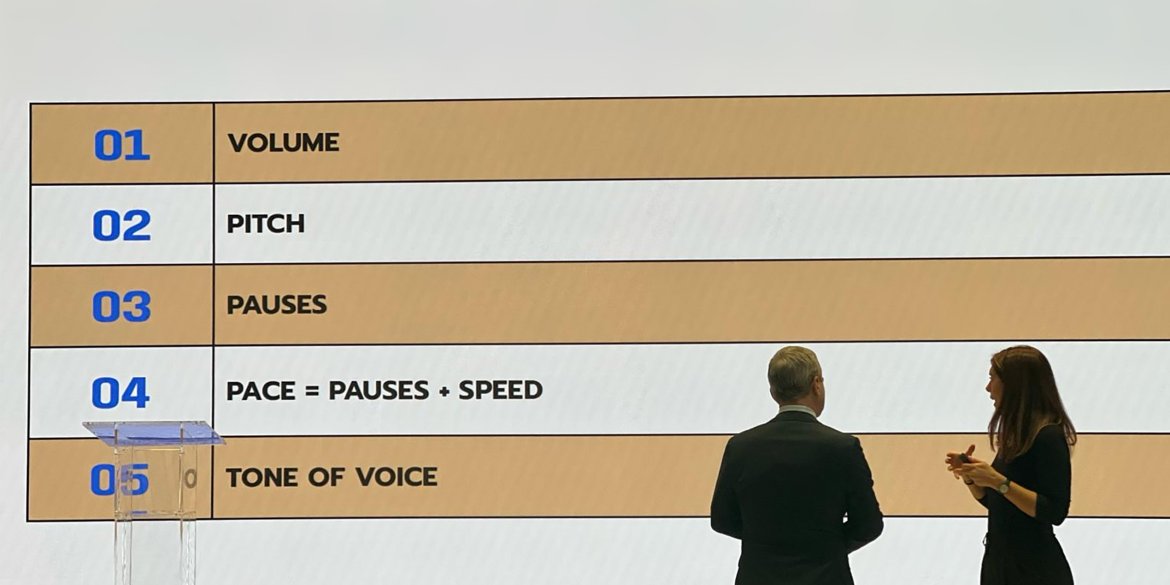 Two presenters discuss vocal delivery techniques in front of a screen highlighting volume, pitch, pauses, pace and tone of voice.