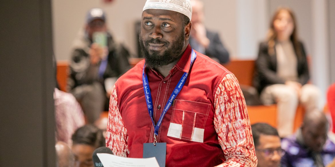 A participant wearing a red shirt and white cap holds a document while speaking into a microphone during a group session.