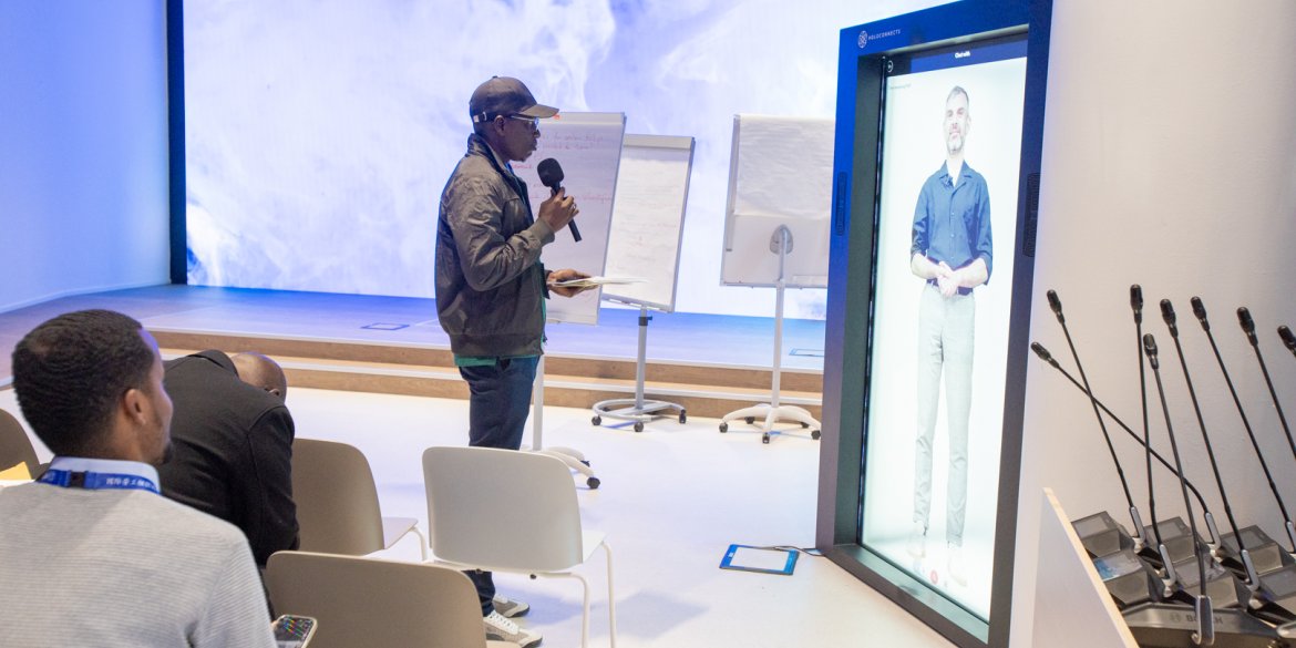 A participant speaks into a microphone beside a life-sized digital avatar displayed on a screen, while others watch.