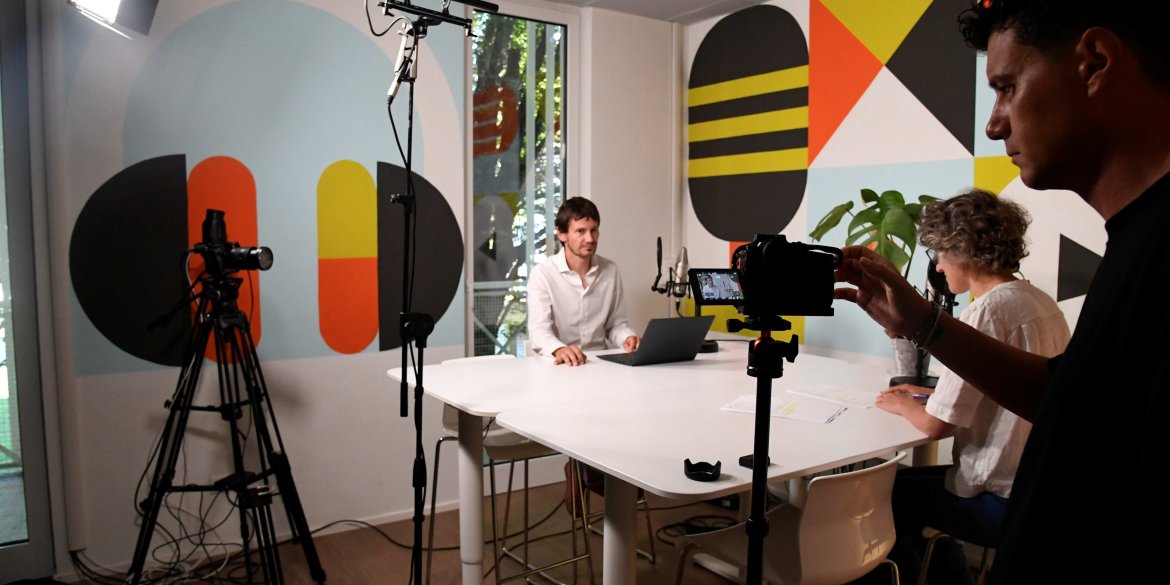 A behind-the-scenes view of a video production in the ITCILO Media Lab, with one person seated in front of a laptop, another reviewing notes, and a camera operator adjusting filming equipment in a brightly coloured studio.