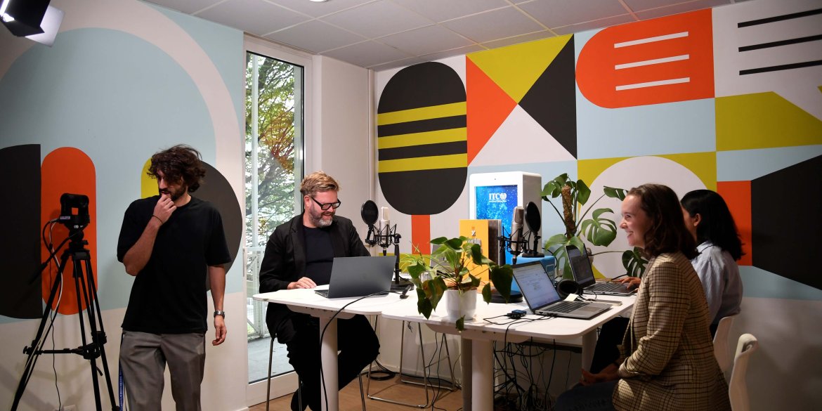 Four people working together in the ITCILO Media Lab, surrounded by laptops, microphones, cameras and studio lighting, against a backdrop of colourful geometric wall murals.