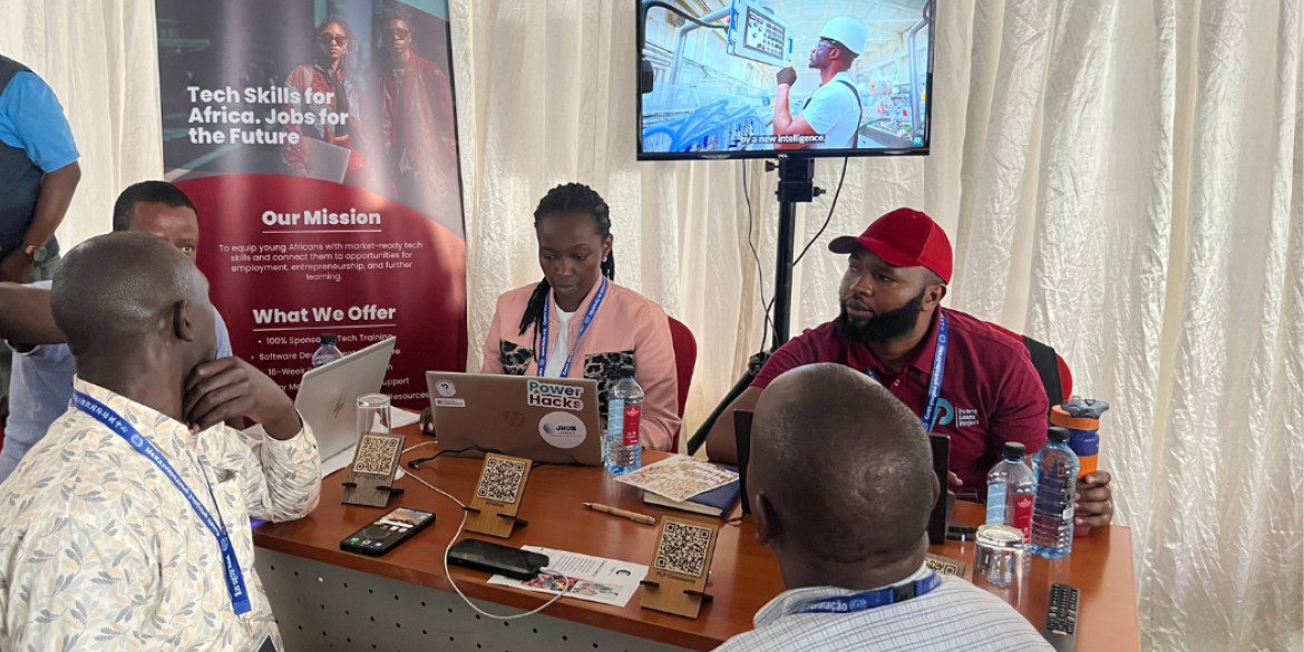 Participants engage in a discussion at a technology skills exhibition stand, with laptops, informational materials and a video display.