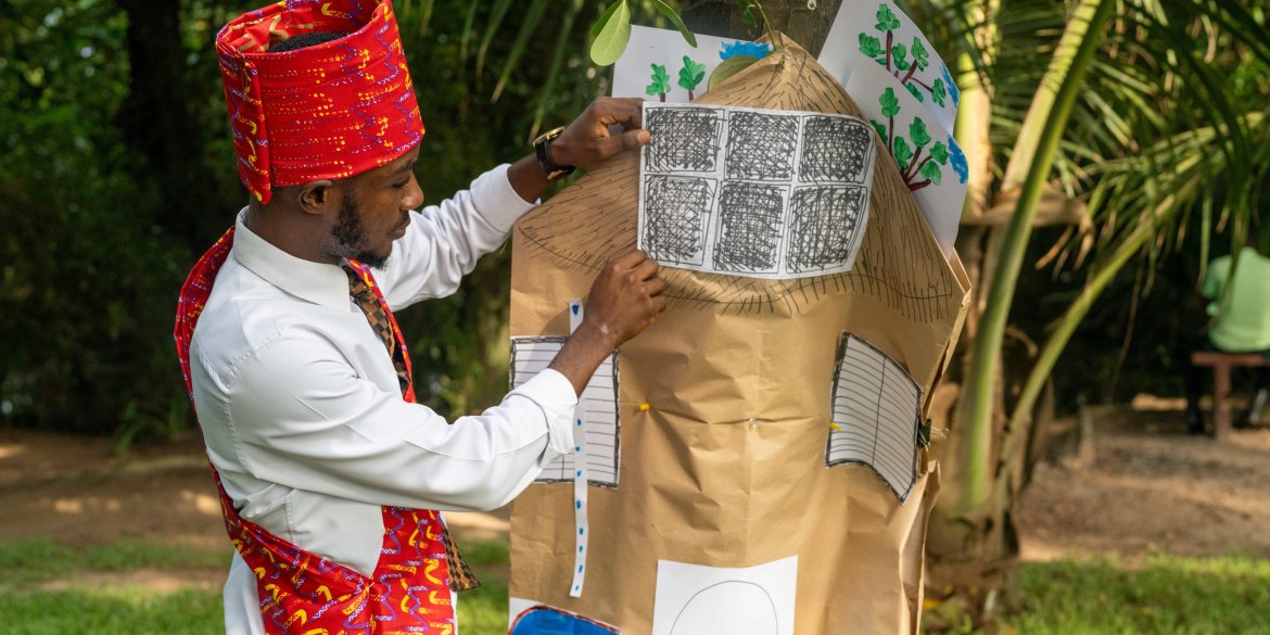 A man in colourful traditional attire presents a handmade model of a house decorated with drawings during an outdoor group activity