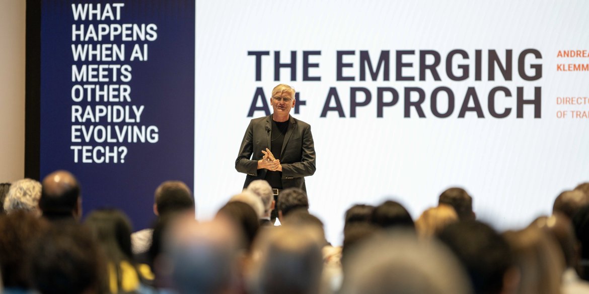 A speaker addresses a seated audience at the ITCILO AI+ conference, standing in front of a large presentation screen displaying “The Emerging AI+ Approach” and a question about AI's intersection with other rapidly evolving technologies.