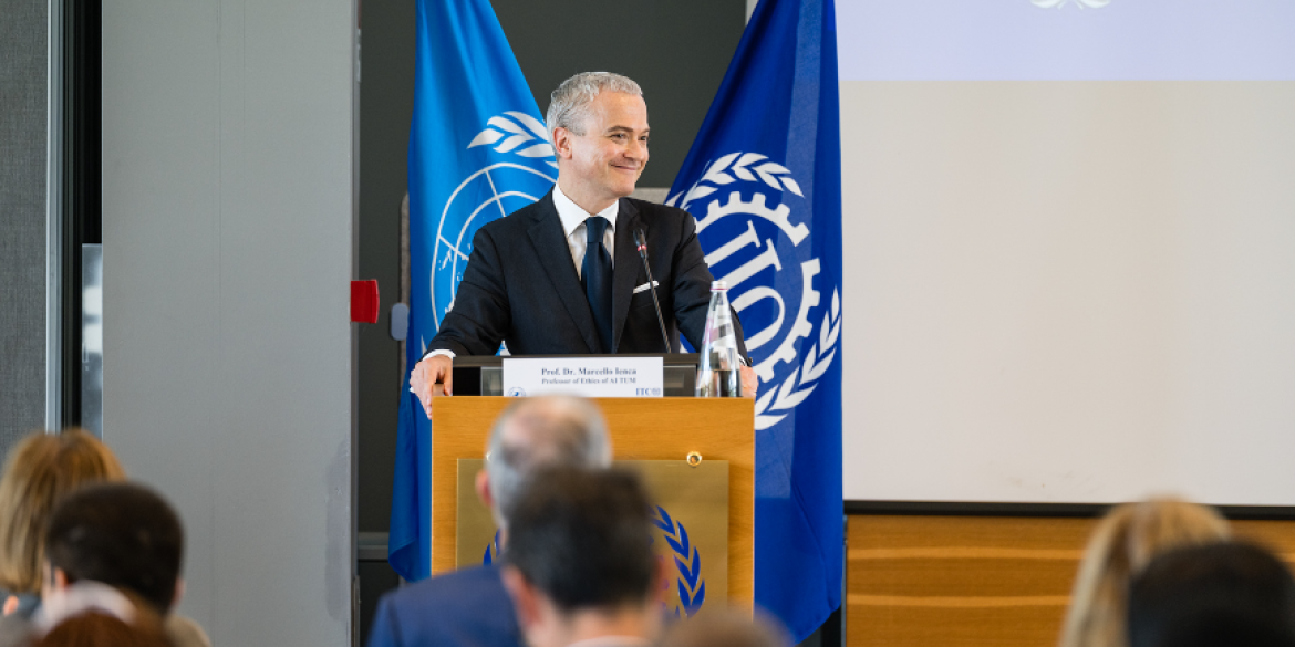 Christophe Perrin, Director of the International Training Centre of the ILO (ITCILO), stands on a stage delivering opening remarks at an event. Behind him are the flags of the International Labour Organization (ILO) and the ITCILO.