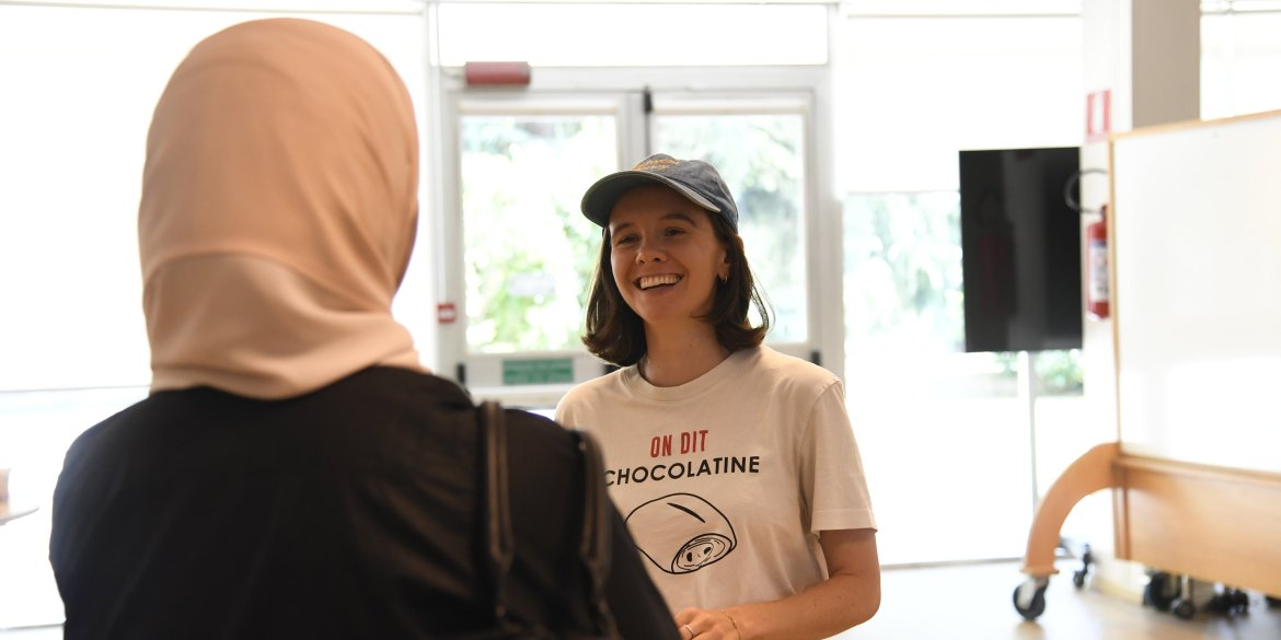 Two people stand talking indoors in a bright meeting space. In the foreground, a person wearing a light pink hijab is seen from behind, facing a smiling person wearing a blue baseball cap and a white T-shirt. The atmosphere is warm, friendly, and conversational.