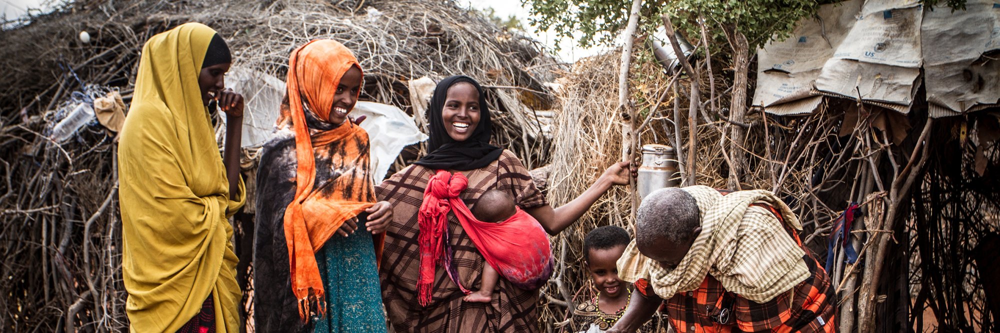 A group of three women, a baby, a toddler and a man gather outside a rustic shelter made of branches and fabric. One woman carries the infant in a bright red cloth sling, while the others wear colorful  yellow and orange headscarves, creating a warm scene of everyday community life.  