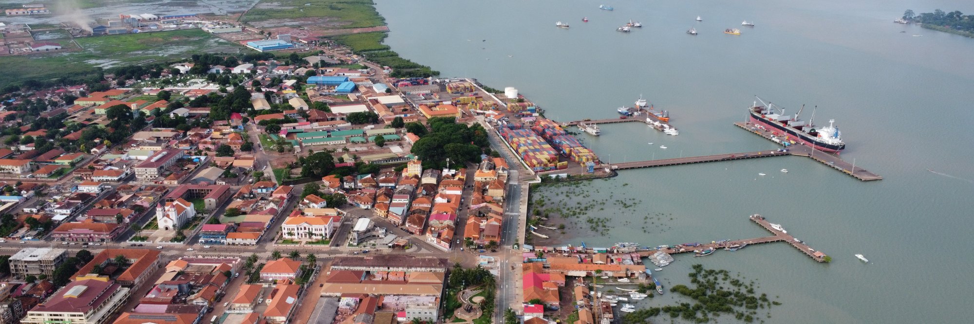 Vista aérea panorâmica de uma cidade portuária cheia de vida, onde os edifícios se encontram com um porto repleto de contentores e longos cais. Os navios de carga repousam na baía tranquila, enquanto mangais exuberantes e zonas húmidas contornam a linha costeira sob um céu enevoado.