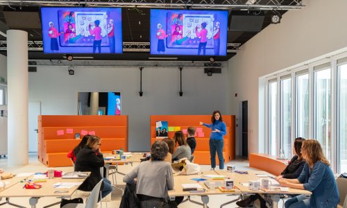 A facilitator leads an interactive workshop with participants seated around tables in a bright, modern training room.