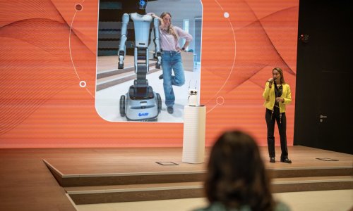 A speaker in a bright yellow blazer demonstrates a small white robot on stage during the ITCILO AI+ Nonference, with a large screen behind her showing a humanoid robot alongside a woman.
