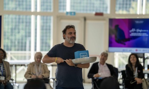 A facilitator stands in the centre of a group of seated participants during the ITCILO AI+ Nonference, holding a frying pan containing a blue box labelled “catch box,” while attendees watch and take notes.
