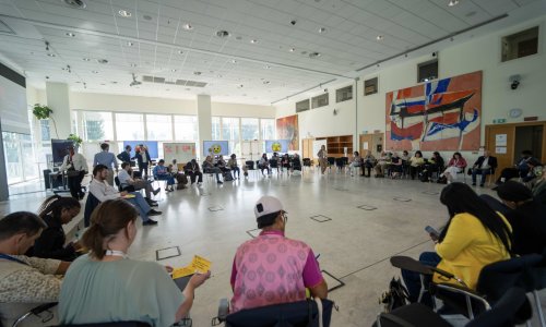 Participants at the ITCILO AI+ Nonference sit in a large circle during an interactive group session, sharing ideas and taking notes in a bright conference hall decorated with colourful artwork.