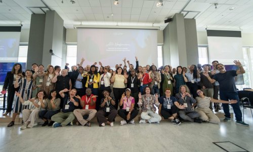 A diverse group of participants and facilitators at the ITCILO AI+ Nonference pose together for a group photo, smiling, waving and raising their hands in a bright conference hall, with the event's branding projected on screens behind them.