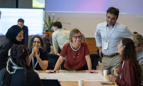 A group of participants at the ITCILO AI+ Nonference engage in a lively workshop discussion around a table, smiling and exchanging ideas over a large worksheet, with other groups collaborating in the background.