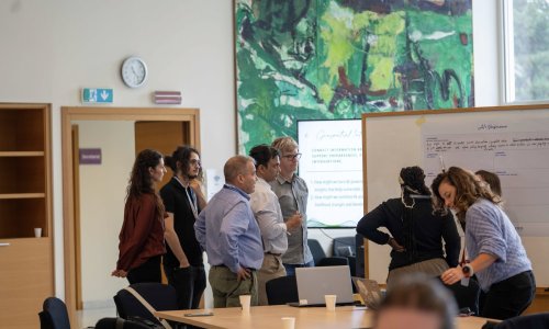 Participants at the ITCILO AI+ Nonference gather around a whiteboard during a collaborative workshop, discussing ideas and reviewing notes on geospatial data and AI, with laptops and presentation screens nearby.