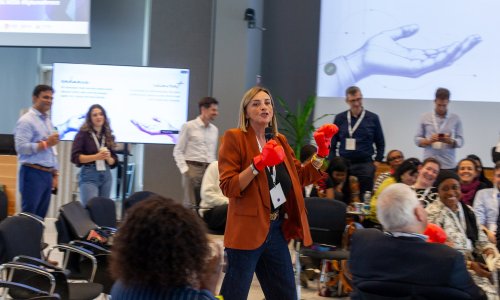 Participants at the ITCILO AI+ Nonference raise their hands during an interactive session, engaging with facilitators at the front of a conference hall.