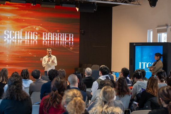 A speaker addressing the audience at the masterclass launch, with a large presentation screen in the background.