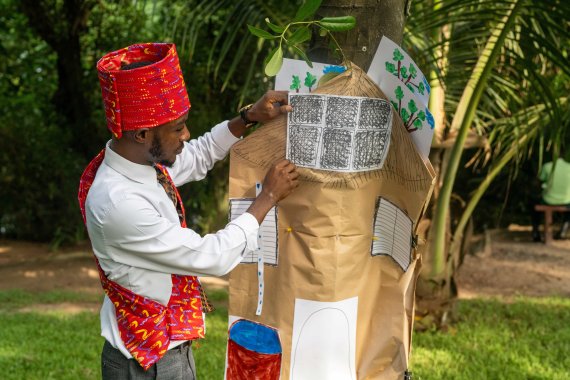 A man in colourful traditional attire presents a handmade model of a house decorated with drawings during an outdoor group activity