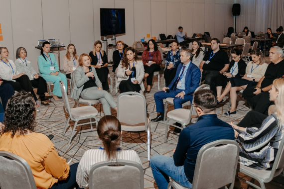 Participants seated in a circle during a group discussion, listening as a woman speaks into a microphone.