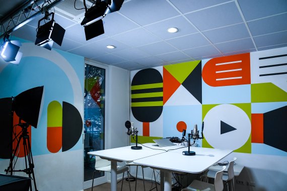 A brightly coloured multimedia recording studio with geometric wall murals, microphones and audio equipment on a white table, overhead studio lighting, and a video camera on a tripod.