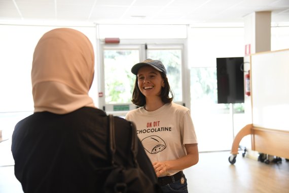 Two people stand talking indoors in a bright meeting space. In the foreground, a person wearing a light pink hijab is seen from behind, facing a smiling person wearing a blue baseball cap and a white T-shirt. The atmosphere is warm, friendly, and conversational.